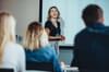 White woman leads a business training in a room full of people of various ages, races, and genders. She is laughing and smiling candidly.