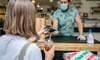 A male food service worker wears gloves and a face mask during the COVID-19 pandemic. He uses a handheld, electronic point-of-sale system to complete a touchless transaction with a female customer using mobile pay to pay for her food.