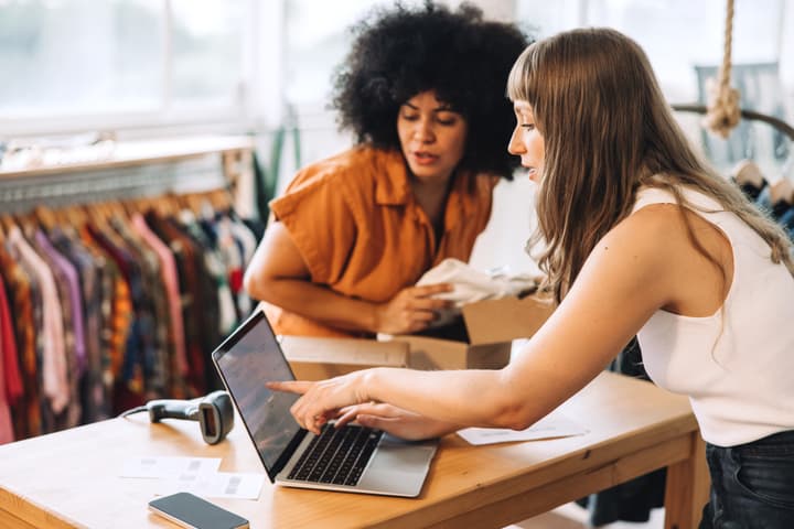 A white woman in a white tank top points to a laptop screen while a black female coworker in a burnt orange button up shirt looks on while unpackaging a small cardboard box.