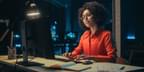 A black woman with glasses sits at computer working for technology company at night