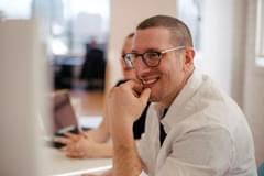 A millennial-aged white male smiles in a modern office setting. He appears to be engaged in a lively conversation with colleagues.