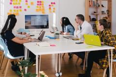 A small group of millennial-aged professionals of various races and genders gather around a modern, bright meeting space with their laptops to work on a project. Photo by Jason Goodman via Unsplash.