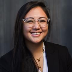 A portrait of Propeller employee Lena Chhay. She wears a white blouse and a black blazer in front of a dark, grey, wooden background. She wears glasses with a clear, acrylic frame.