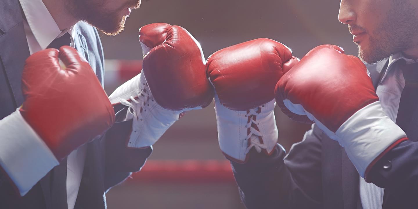 Close up of two business men in suits wearing boxing gloves in a boxing ring. Their gloves are touching.