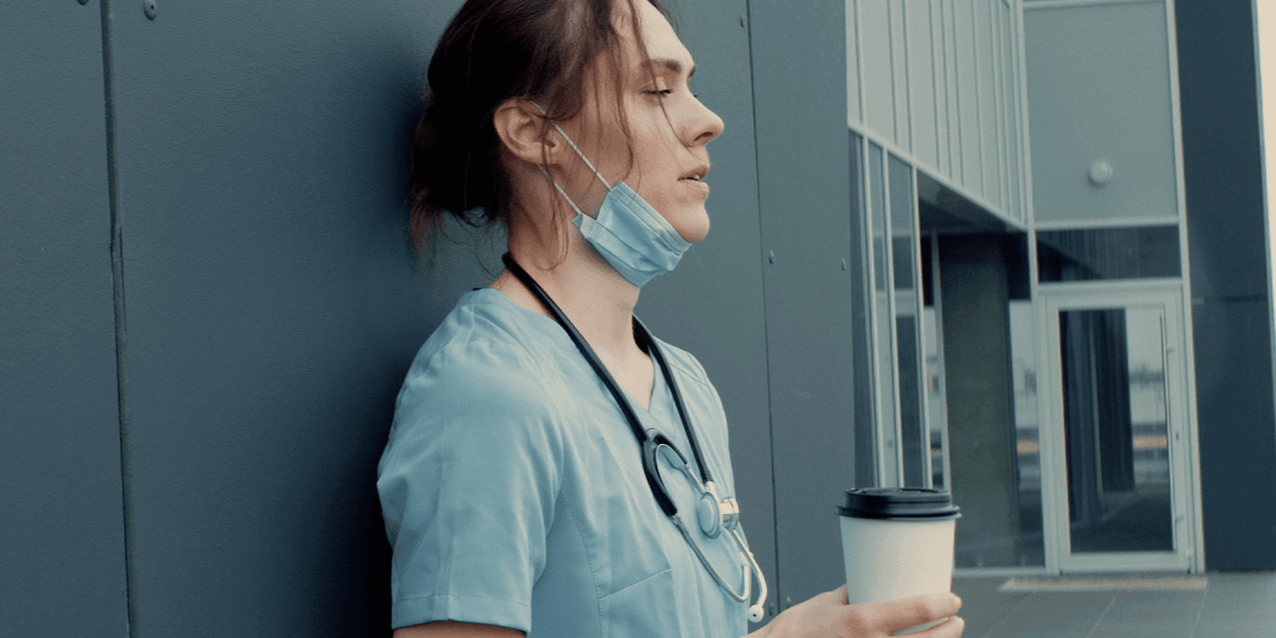 Image of a white, female medical professional wearing pale blue scrubs and a stethoscope. Her face mask is pulled down below her chin and she is holding a to-go cup of coffee. She appears exhausted.